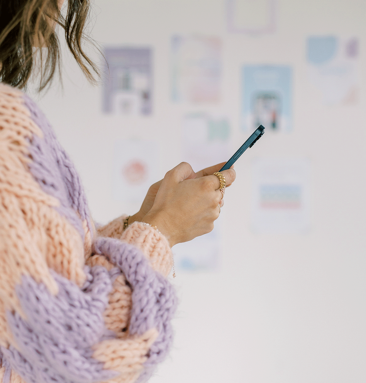 Frau in Strickpullover hält Smartphone in den Händen, vor einer Wand mit sanften Farben und Inspirationen.